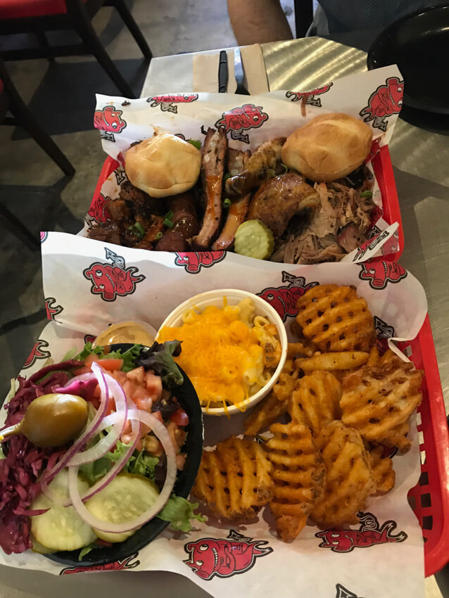 Two paper-lined baskets filled with cooked chicken, beef, bread rolls, a bowl of salad, a cup of mac and cheese, and waffle fries