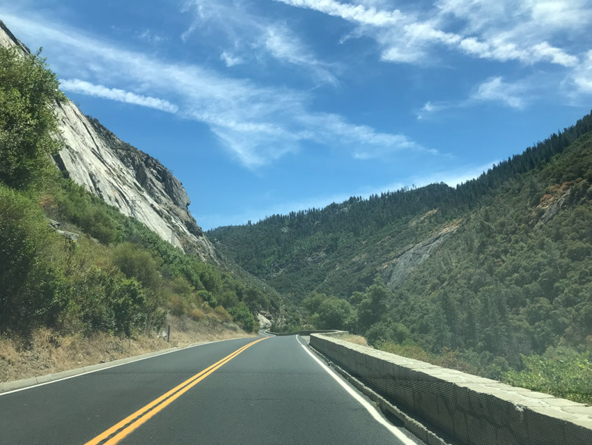 A road with a shallow wall to the right, granite rock to the left and green shrubbery on the mountains in the distance