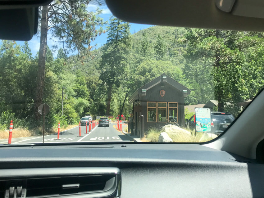 A view from inside a car of a national park with many trees. There are some cars up ahead and a small building where a woman in uniform is standing outside. Several stop signs can be seen and the word STOP is painted on the road.