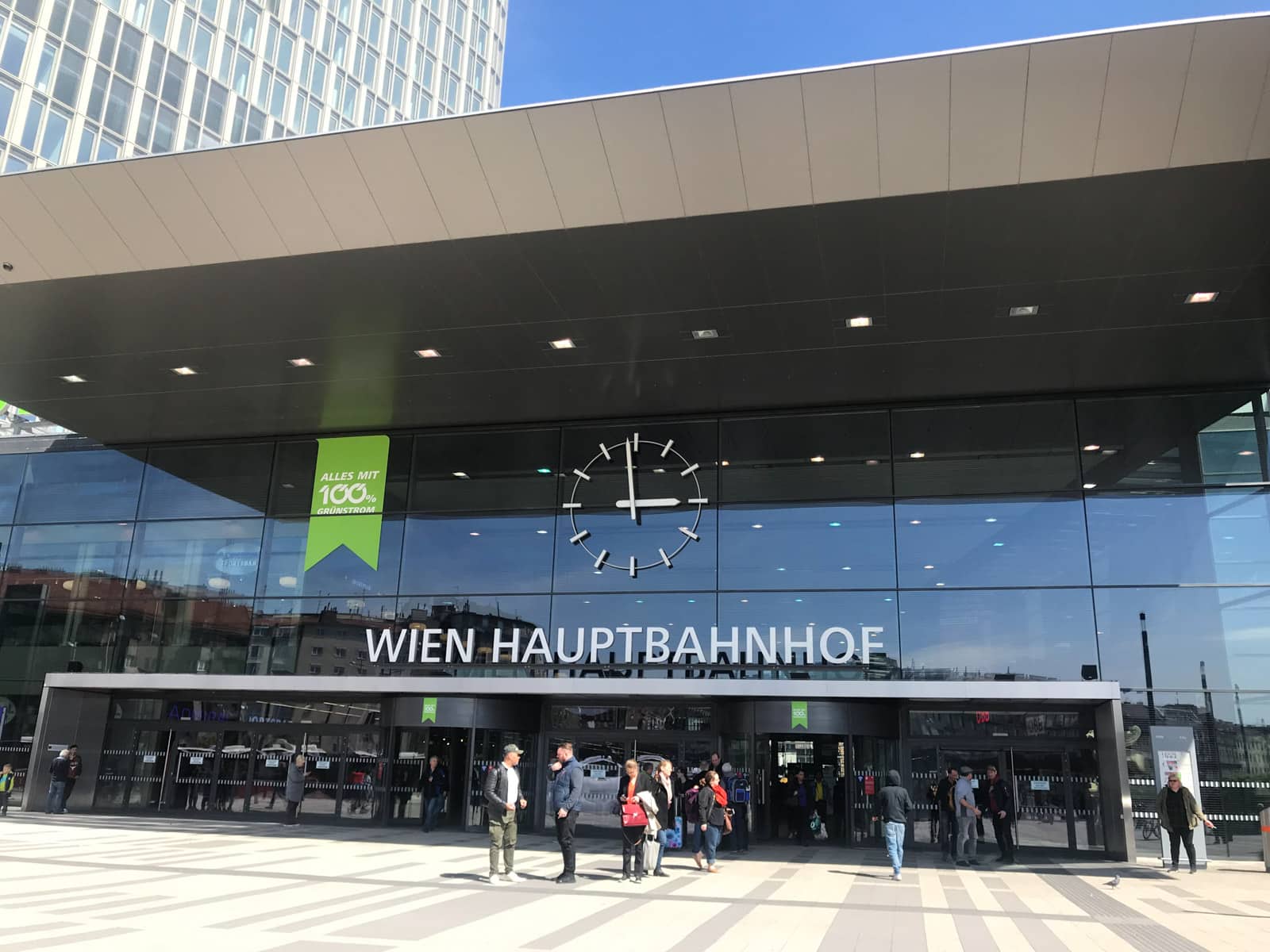 The entrance of a building with the sign “Wien Hauptbanhof” above the glass doors. An analog clock on the building face reads that it is almost 3:00pm