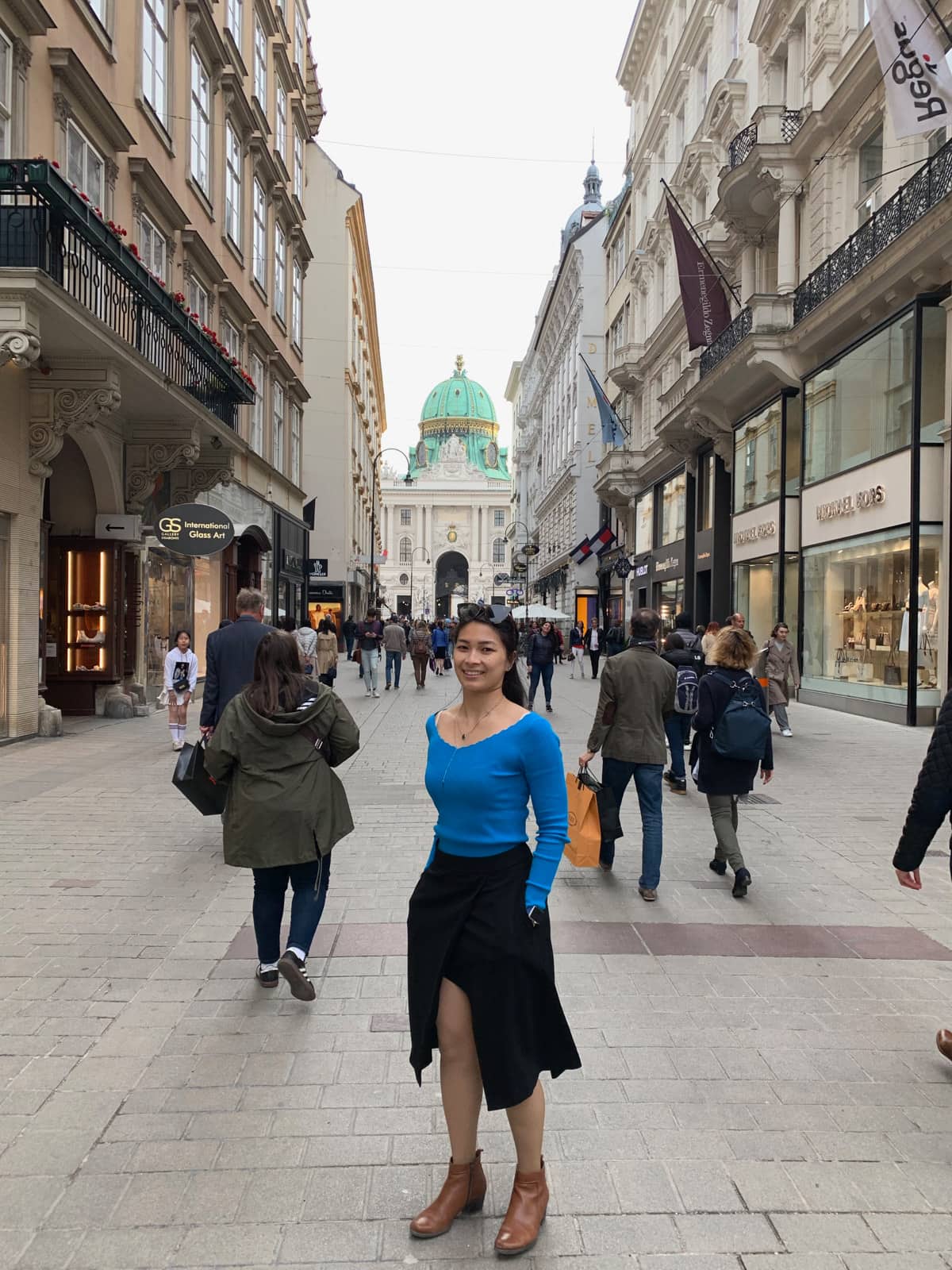 A woman wearing a blue long sleeved top and a long black skirt. She has her hands in her pockets. She’s standing in a pedestrianised open street mall and in the background is an old building with a green dome-shaped top