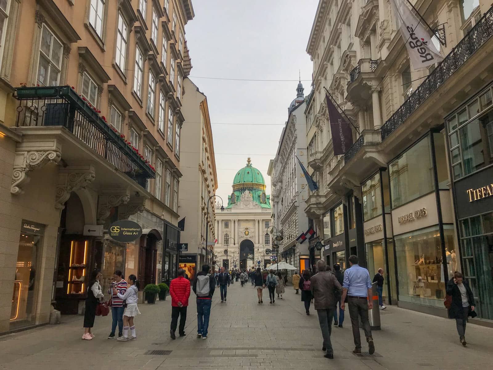 An outdoor shopping mall with shopfronts to the sides, and people walking and browsing. Ahead, centre of frame, is an old building with a green dome top