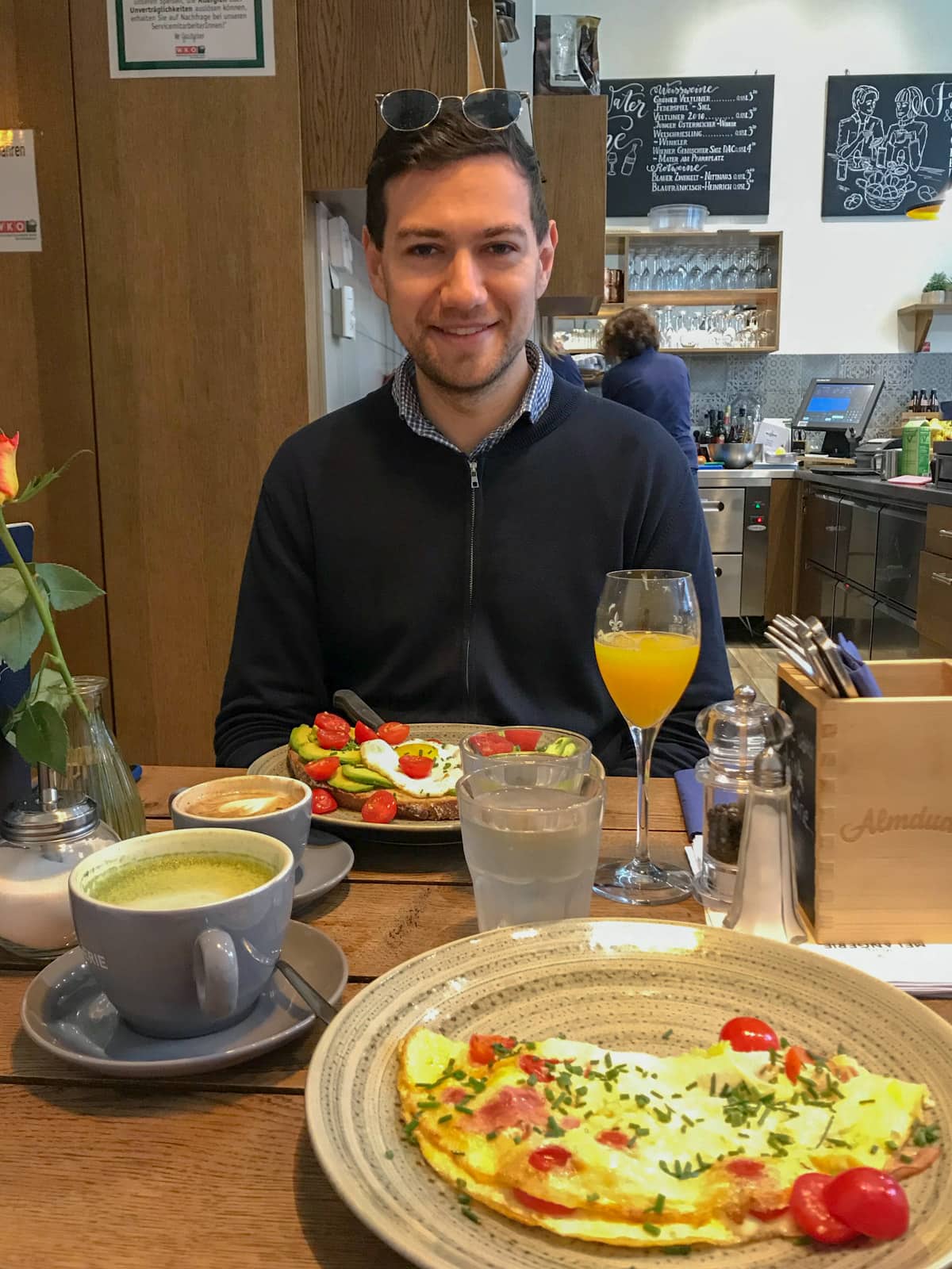 A man smiling, sitting at a table which has breakfast served. There are two plates, one with an omelette and the other with avocado on toast. There is orange juice in a glass and a couple of coffees on the table