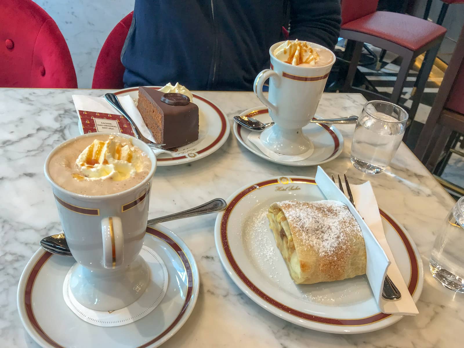 A marble table set with plates of apple pie and a slice of chocolate cake, and two tall mugs of coffee with caramel sauce and whipped cream