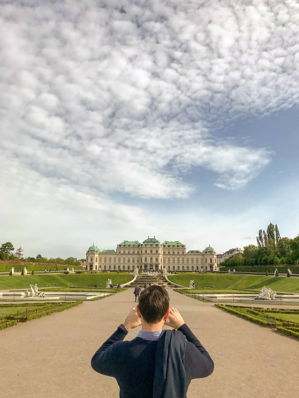 An old palace with a wide path leading up to it. In the foreground a man is taking a photo using his phone.