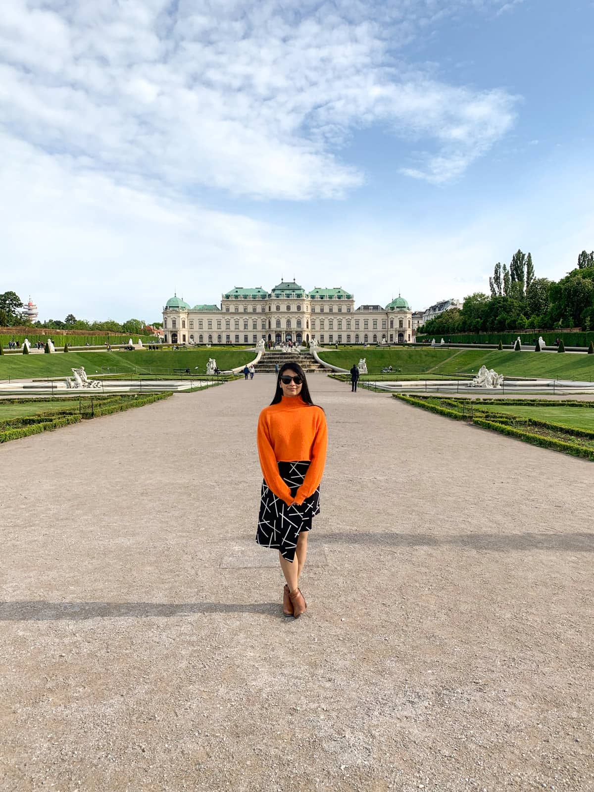 A woman wearing sunglasses, with an orange sweater and a black and white patterned skirt, standing in the middle of a wide path. The path leads to a palace in the distance, with green roofs. Beautiful gardens make up part of the grounds