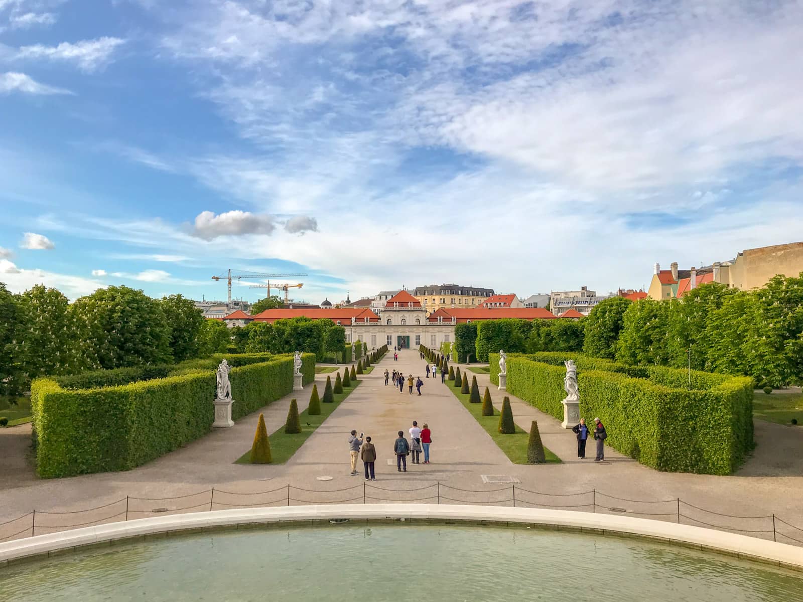 The grounds of a palace with some very neatly trimmed green hedges to the sides, and a path leading up to a building. There are some statues near the hedges. In the foreground is a large man-made pond