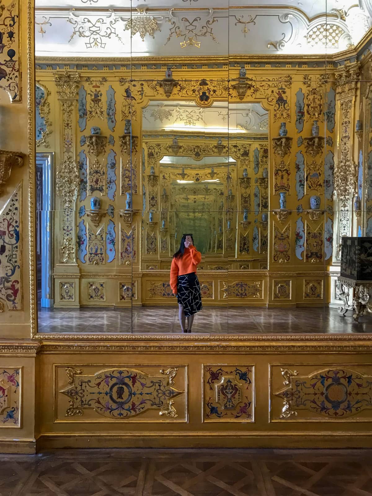 A mirror reflection of a woman taking a selfie using her phone. She is wearing a layered patterned black skirt and an orange sweater. Her face cannot be seen