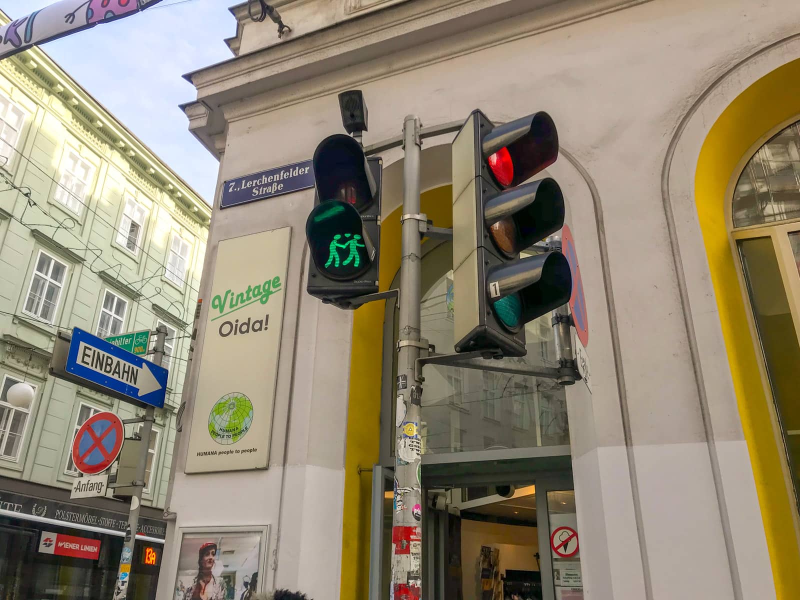 The pedestrian lights on a street corner, lit up green, with the image showing two figures walking and holding hands. In the background is a vintage clothing store.