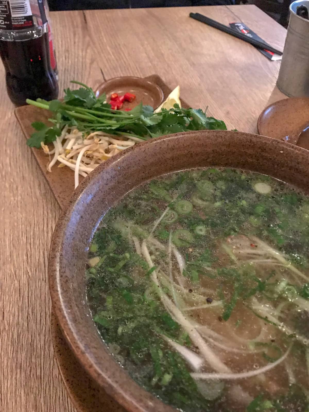 A brown bowl of Vietnamese pho noodles. In the background is a small plate with bean sprouts, coriander and chilli