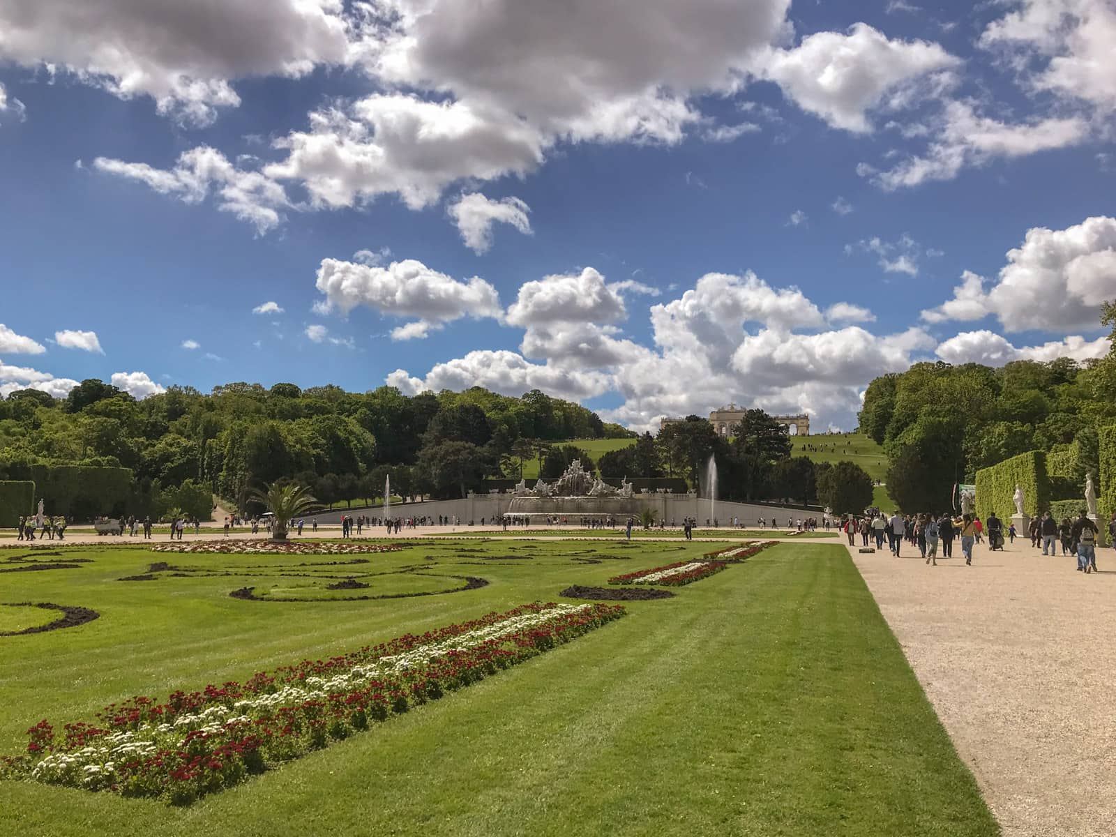 The grounds of a large garden, with wide walking paths to the sides of the mowed grass. The grass has flower arrangements in straight lines, and some swirly patterns dug into the grass to reveal the dirt below. Beyond the garden is a fountain and there are people walking on the paths.