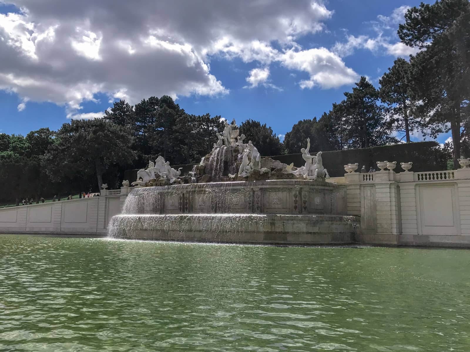 A large fountain with sculptures, water going into the man-made pond below. There are trees in the background and the sky is bright blue.