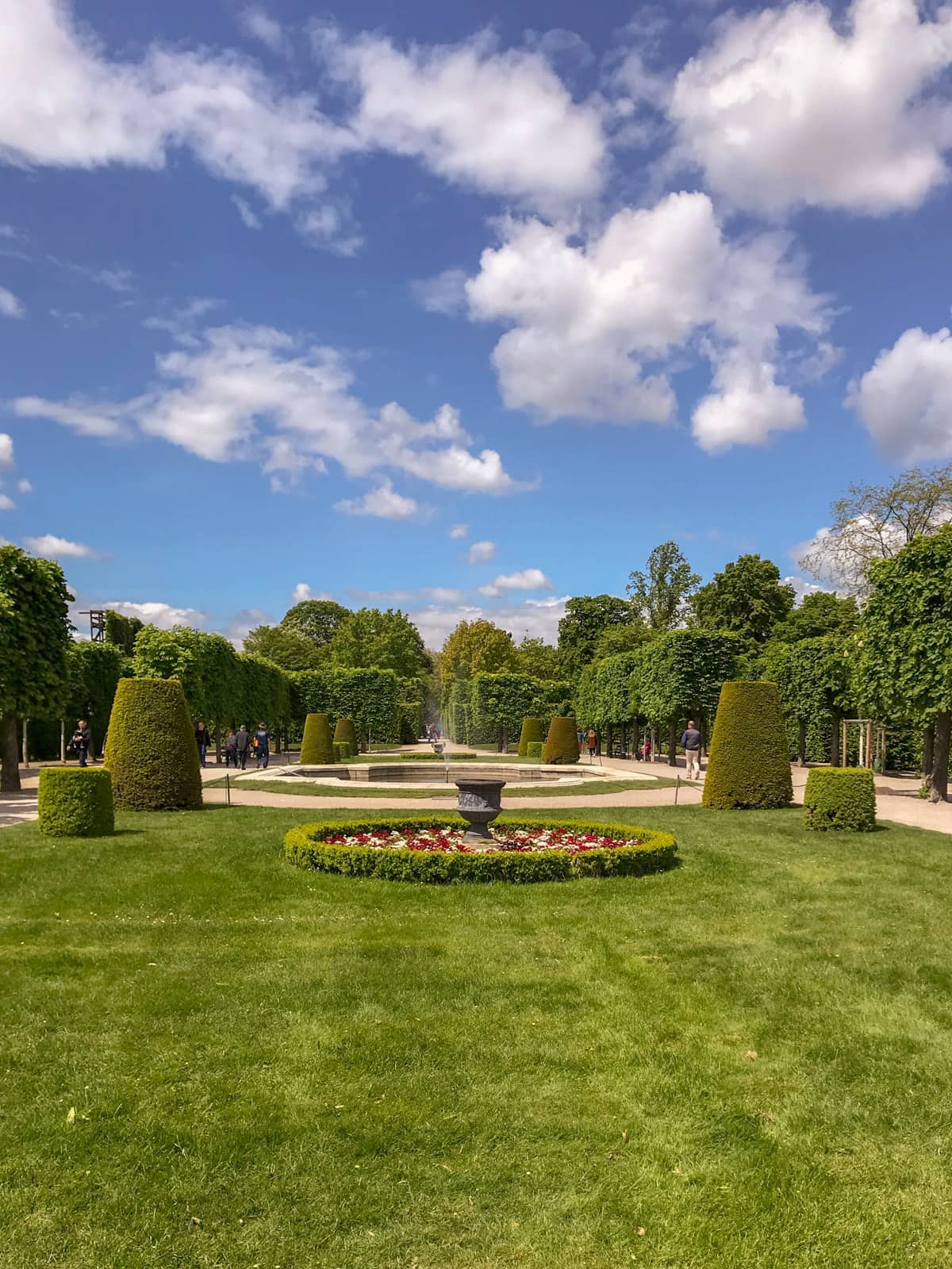 A freshly mowed garden with some hedges to the sides of the garden paths. A small water feature can be seen in the distance, and an arrangement of flowers in the centre of frame. The sky is bright blue