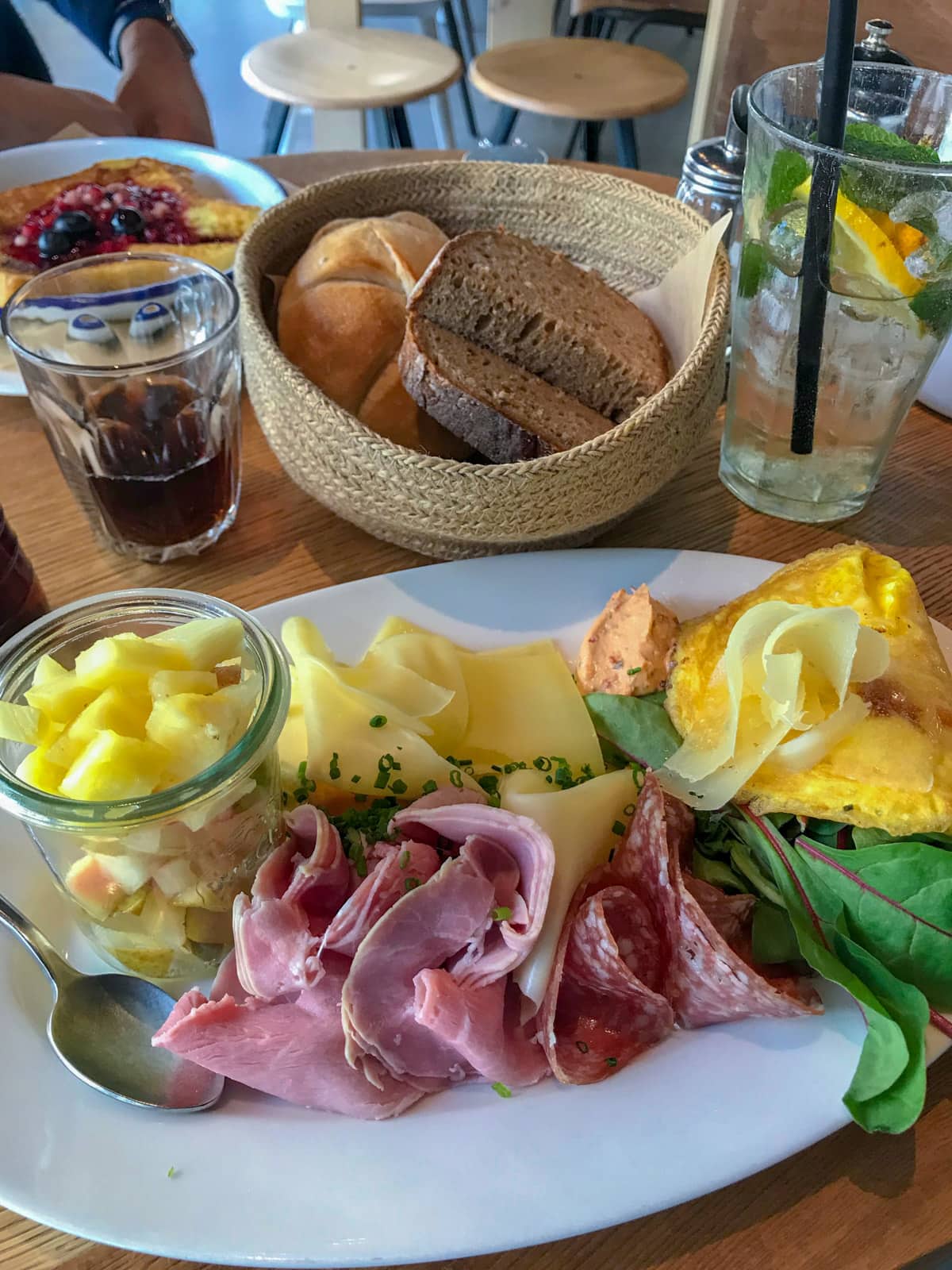 An oval-shaped plate on a table, on which is served some cold cuts of meat, cheese, eggs, and a cup of fruit. In the background is a glass of Coca Cola and a small basket of bread