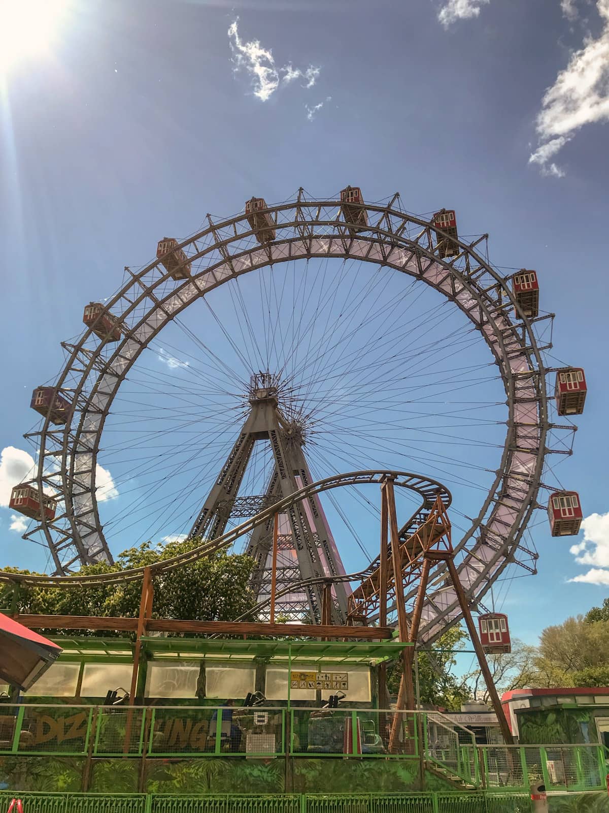 A very old ferris wheel, seen during the day. In front of the ferris wheel is a mouse-trap style ride slightly obstructing the view