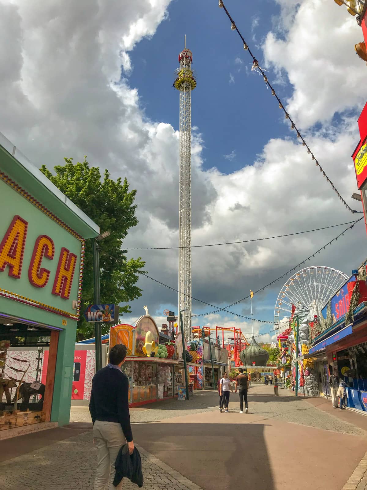 An amusement park during the day, with cables of lights across the way. There is a man in a dark jacket in the foreground, looking up at an attraction that is a tower with passengers experiencing a drop from the great height