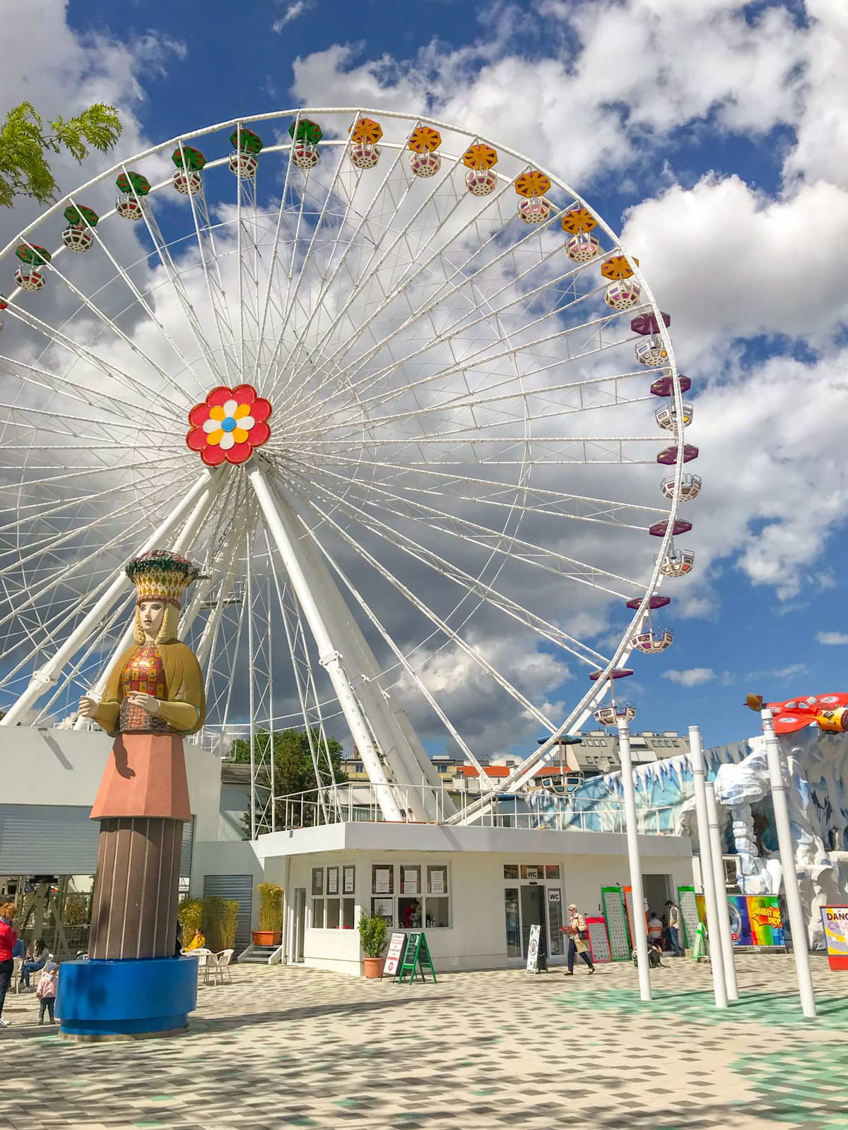 A giant white ferris wheel with bright blue sky in the background. The sky has many clouds. In the foreground is a large colourful figurine with a patterned hat