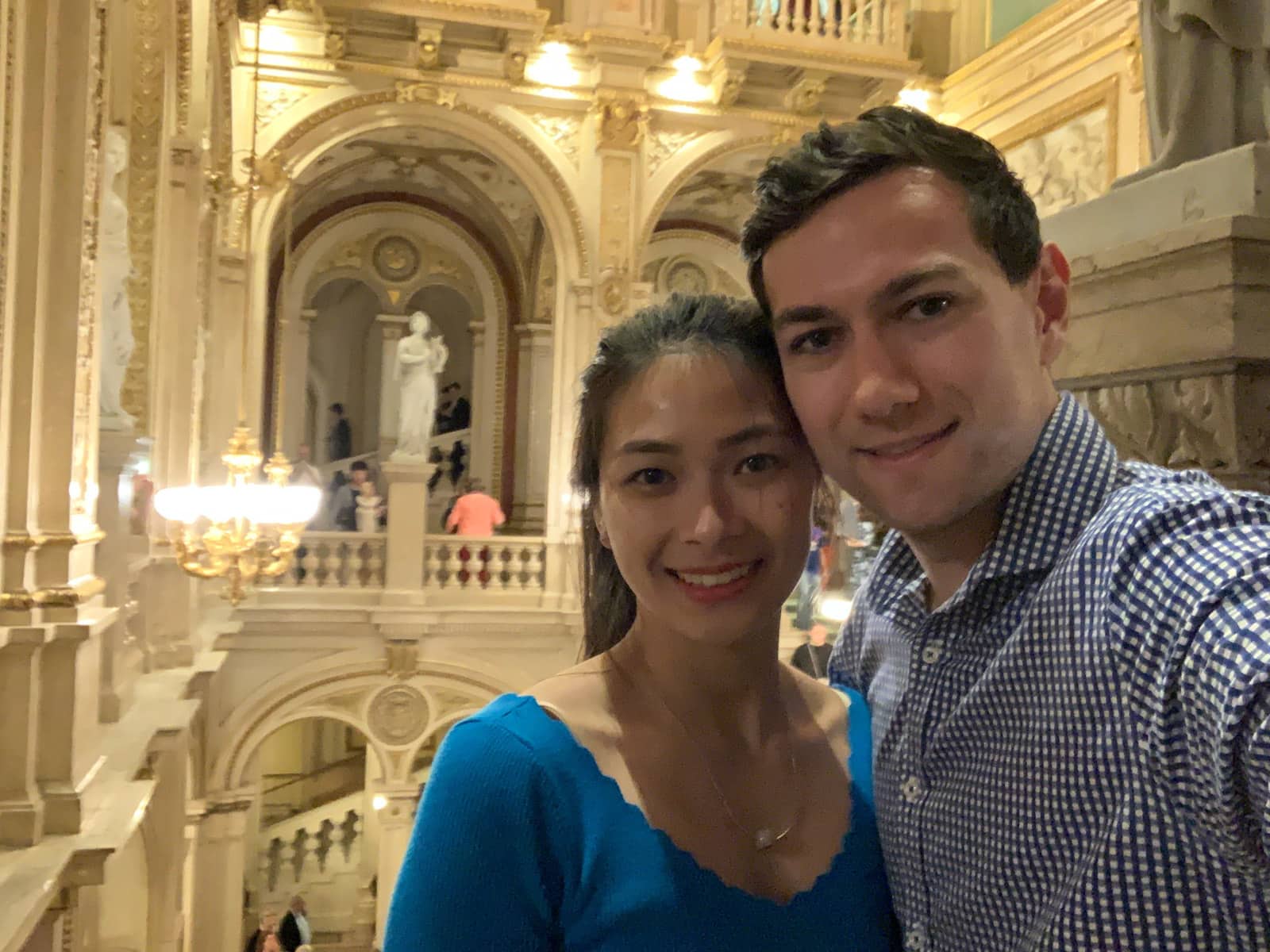 A selfie of a man and woman smiling. The woman is wearing a bright blue top and the man is wearing a checkered collared shirt. They are in an old theatre building.