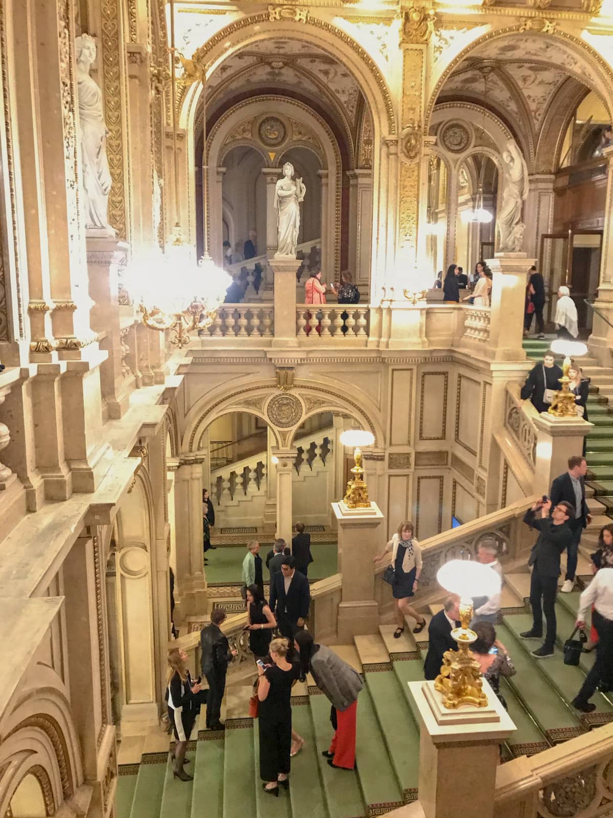 The inside green carpeted stairs of an old building, seen from an internal balcony. There are people on the stairs dressed up and taking photos of each other.