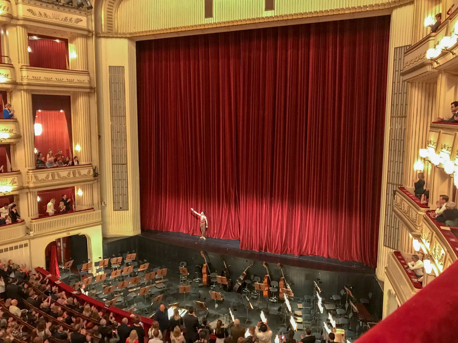 A theatre seen from a box seat. It is the end of a show, and a red curtain is up on the stage. There is one performer taking a bow. The orchestra area is empty.