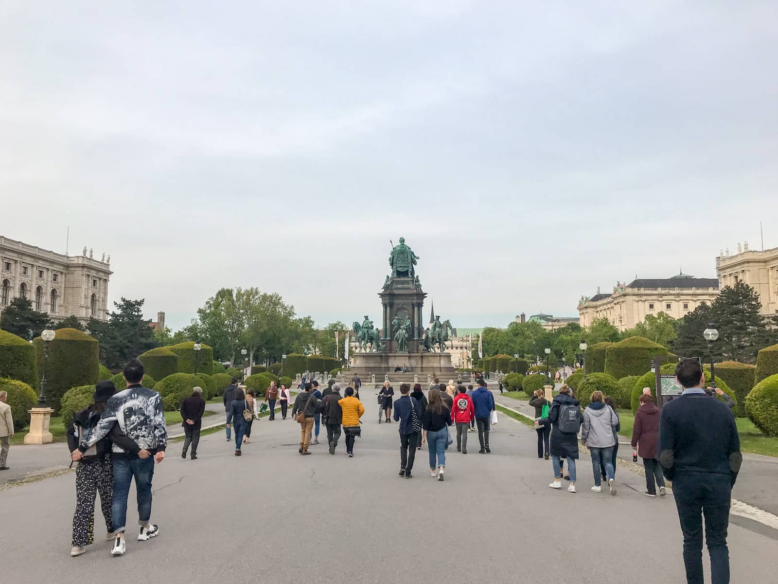 An open path with a large statue up ahead. Many people are walking down the path, and there are old buildings to the sides