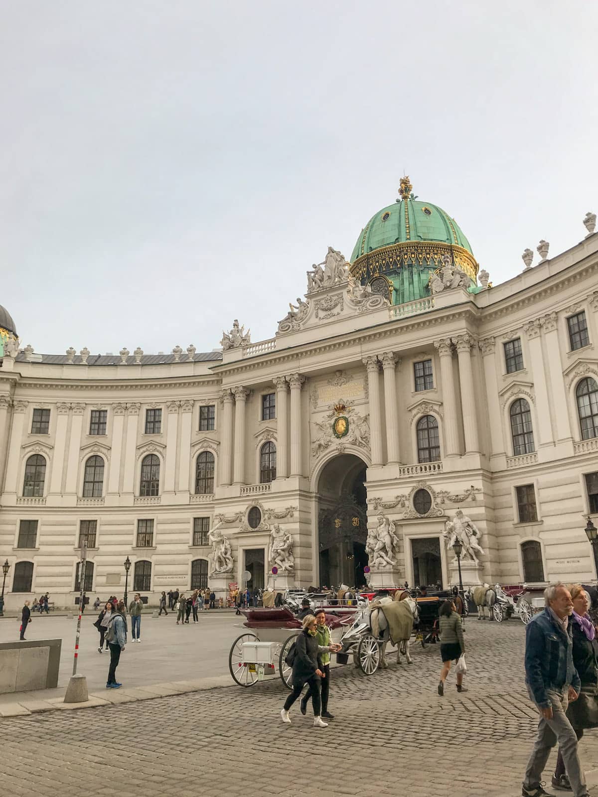 An old building with arched windows and a green dome marking the centre of the building. There is a road that goes through an arch through the building. There is a horse-drawn carriage in the foreground and some people walking around.