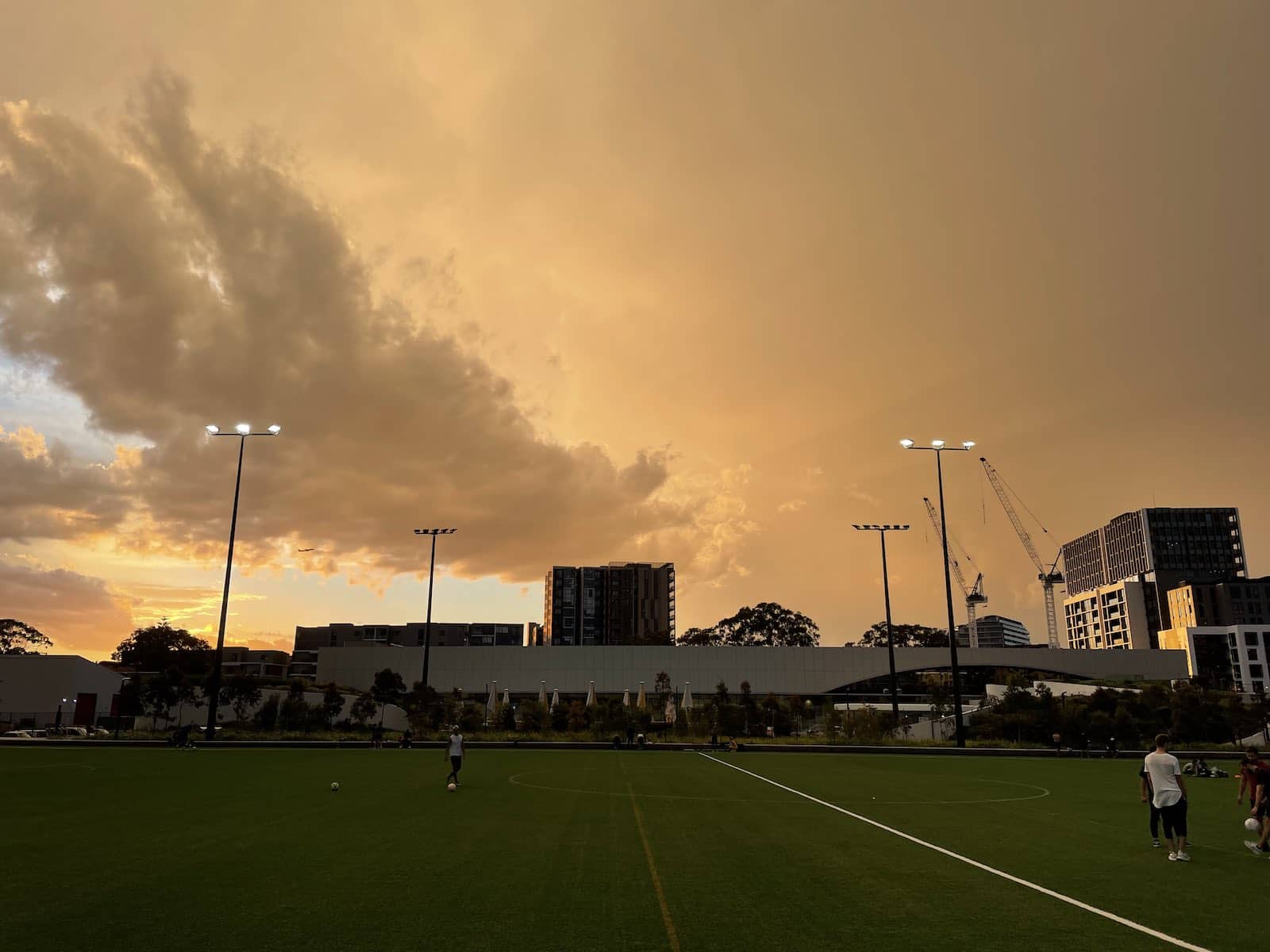 A soccer field at dusk showing a yellow sky with lots of clouds. There are some modern apartment buildings behind the soccer field and some construction cranes in the background.