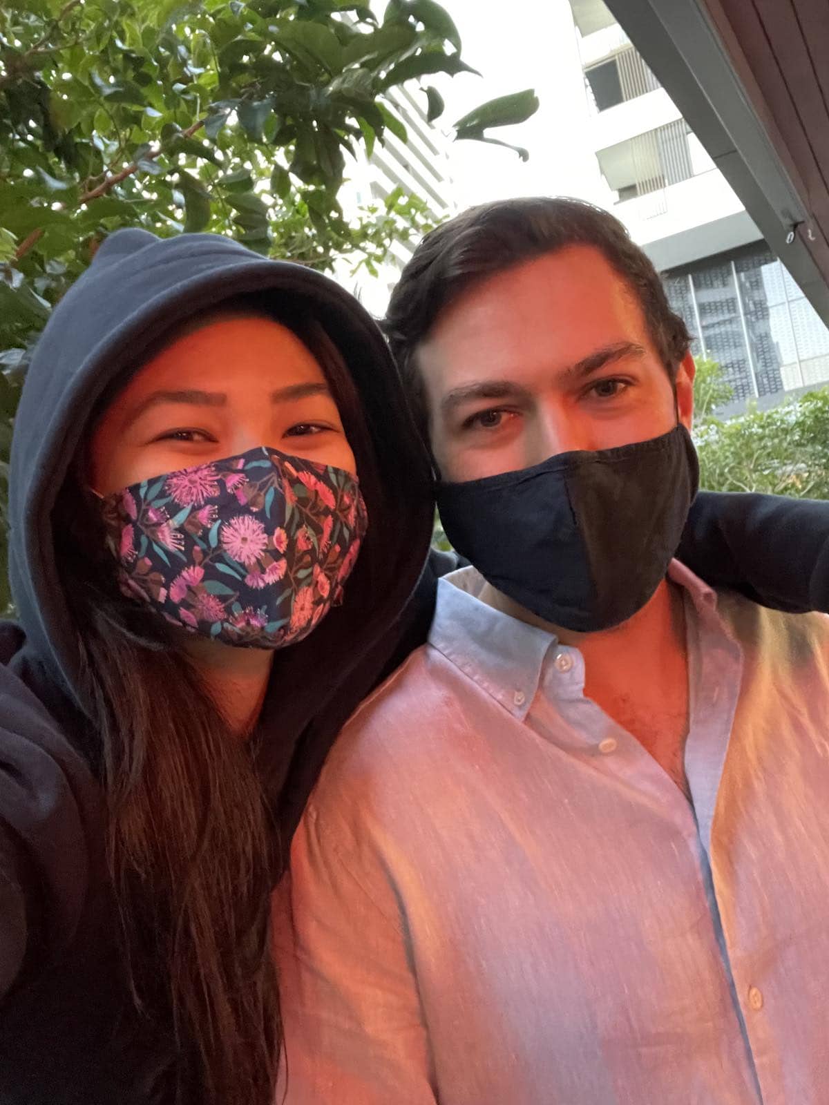 A selfie of a man and a woman, with the woman’s arm around the man’s shoulder. They both have face masks on. The woman has long dark hair and is wearing a navy hoodie and her face mask has a bright gumtree pattern on it. The man has dark hair, a have mask, and is wearing a linen shirt. There is warm red lighting projecting a slight redness on their faces.
