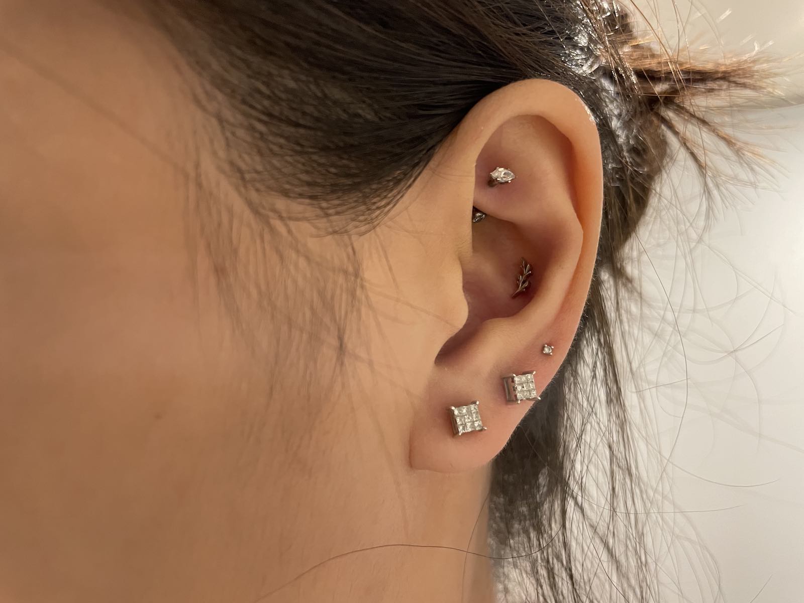 A close-up of a woman’s ear. She has three lobe piercings, a rook piercing and a conch piercing. The lobes have studs in them with clear gems, the conch piercing has a leaf-shaped stud, and the rook has a stud and a crystal
