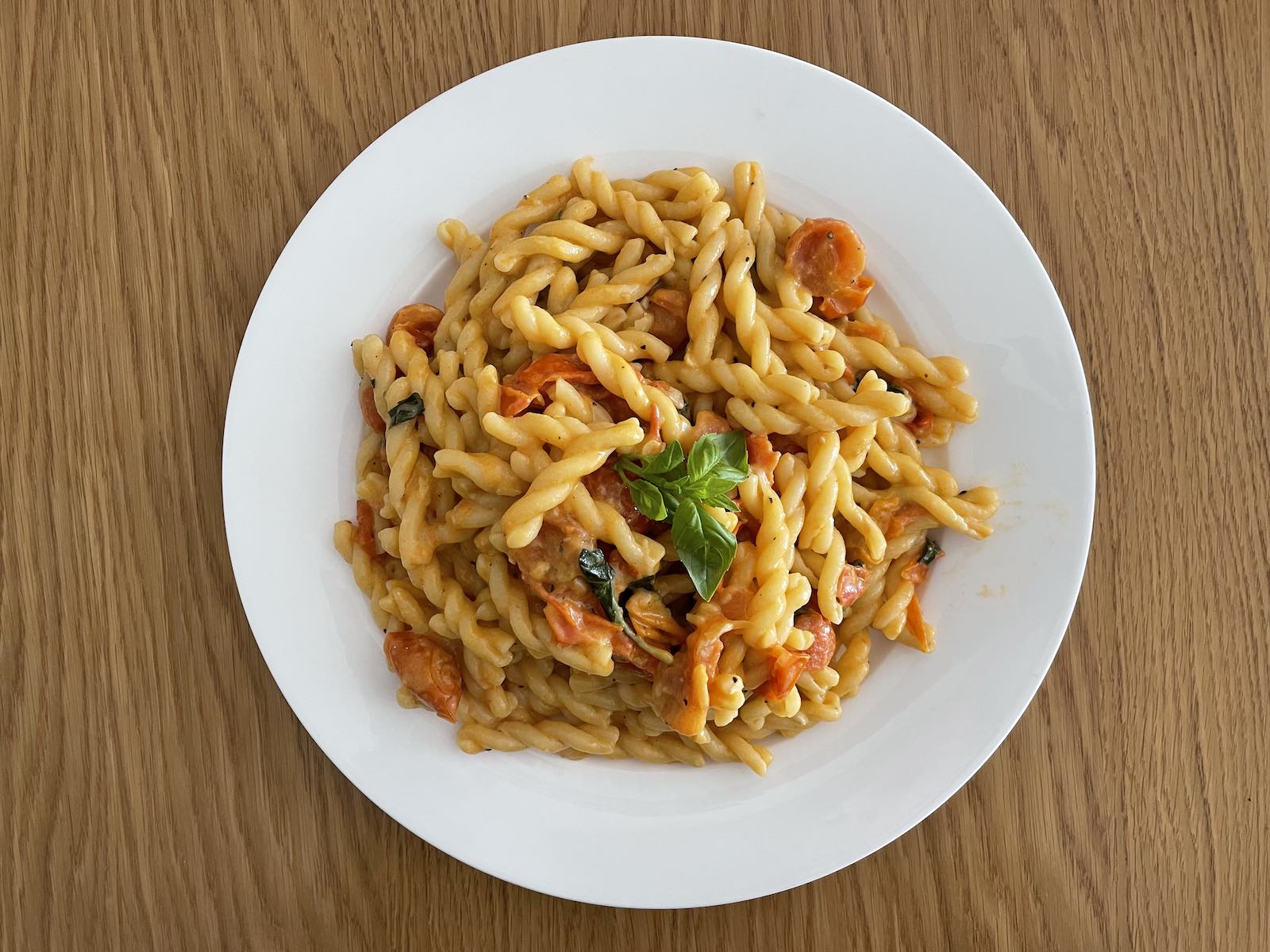 A white plate of curly pasta on a wooden table. The pasta is cooked with cherry tomatoes, basil, and has a slightly creamy texture.