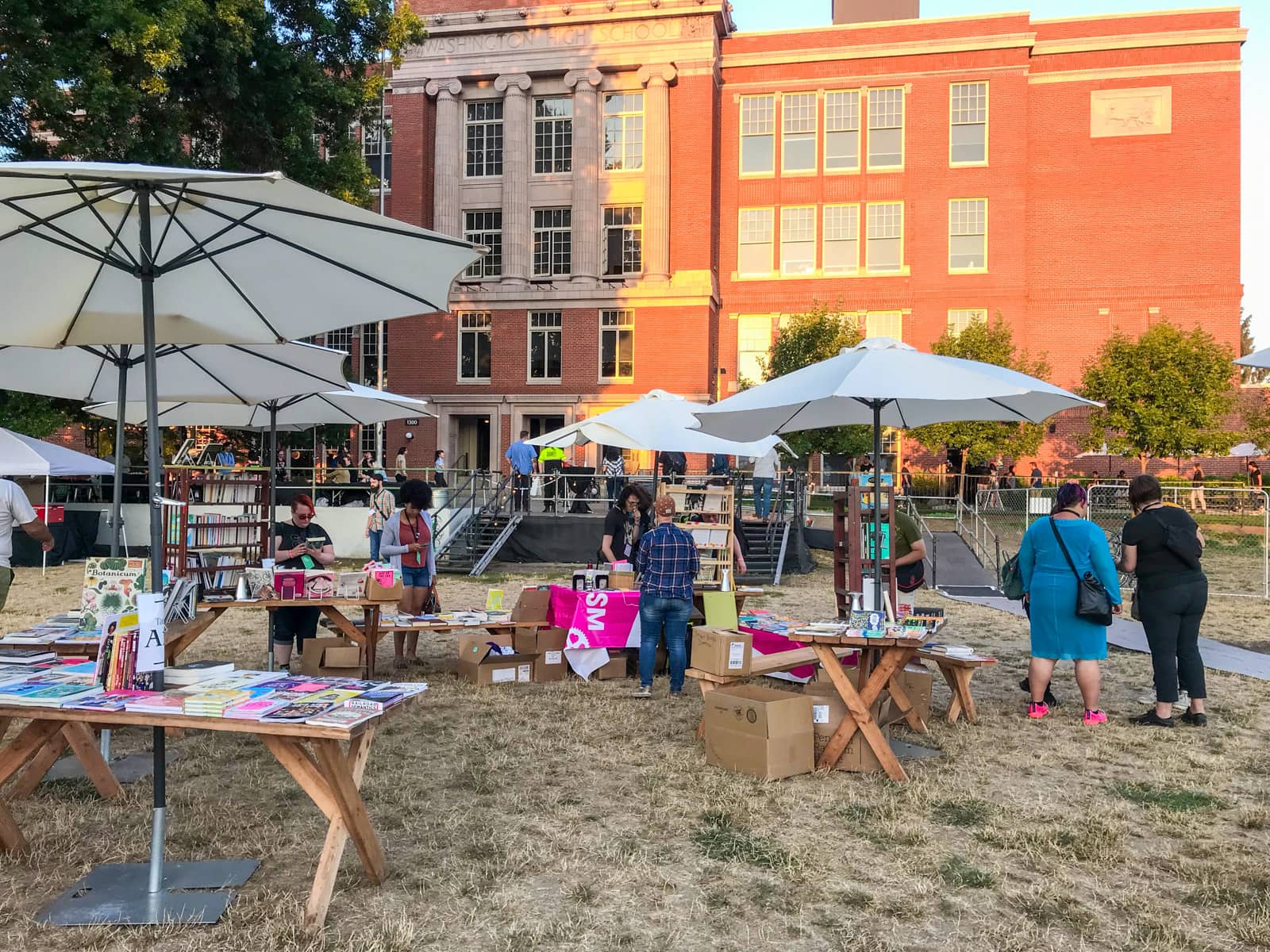 An open park area with stalls set up to sell books. There are some umbrellas providing shade, and the direction of the sun indicates it is the afternoon.