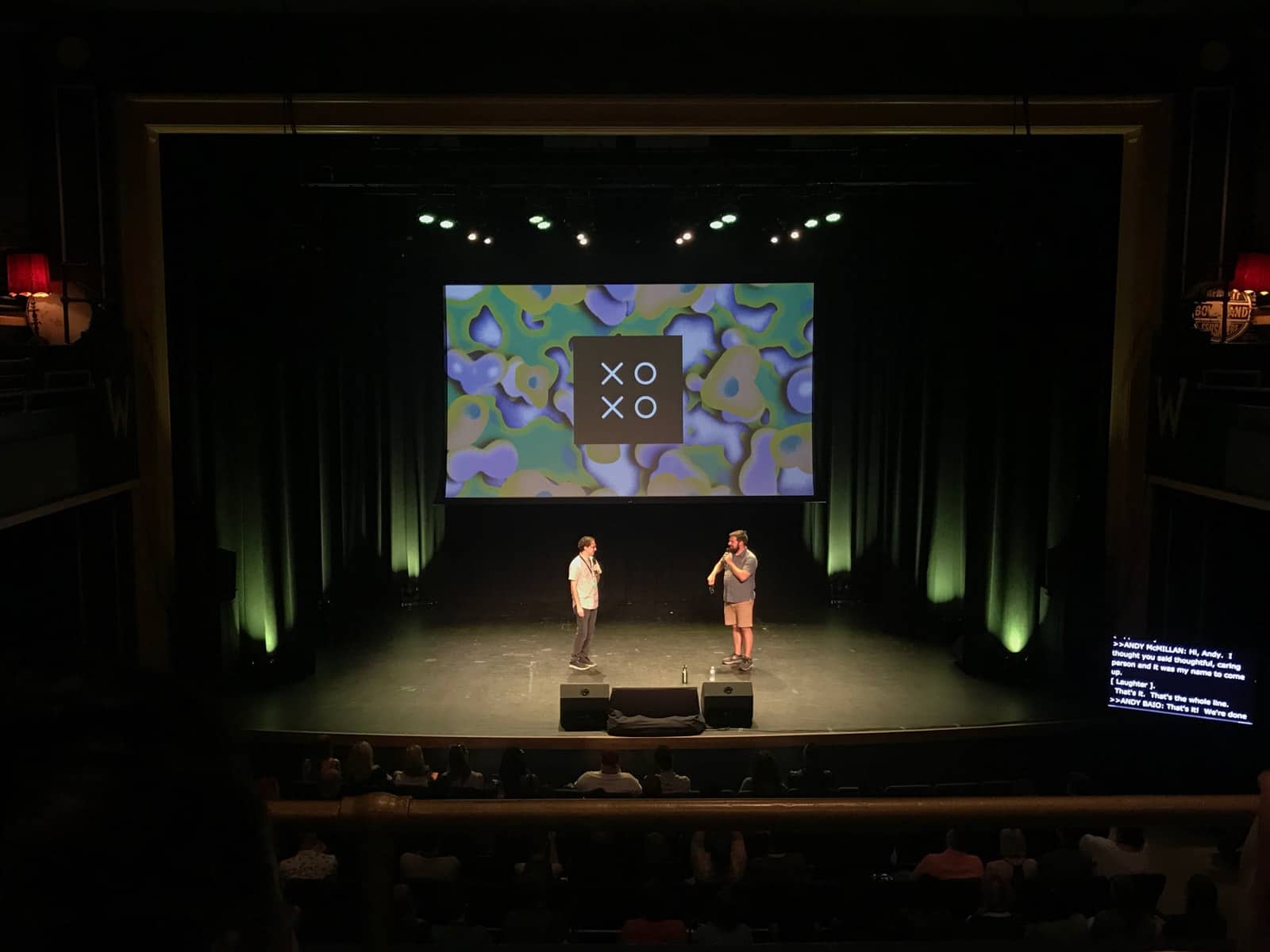 A stage with two men standing on it, holding microphones, in front of a screen with a coloured pattern and the text “XOXO”