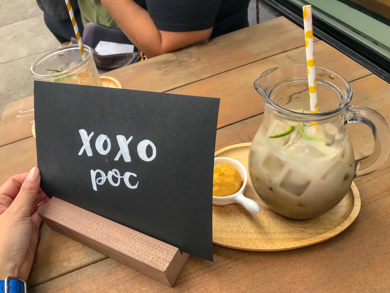 A glass jug of hojicha milk tea served on a tray, alongside a small black cardboard sign reading “xoxo POC”