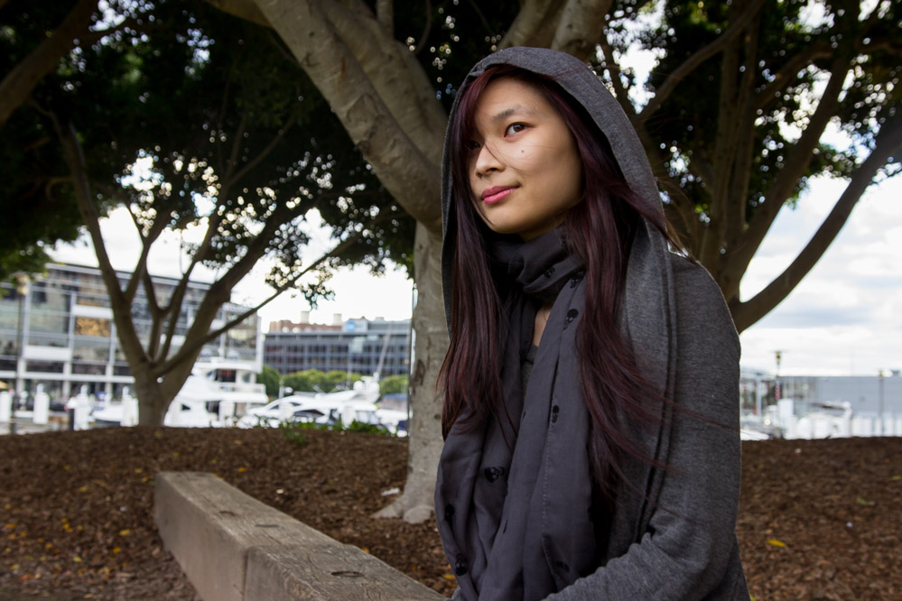 A girl with long dark purple hair, with a grey hood on. She is wearing a grey scarf with skulls on it.