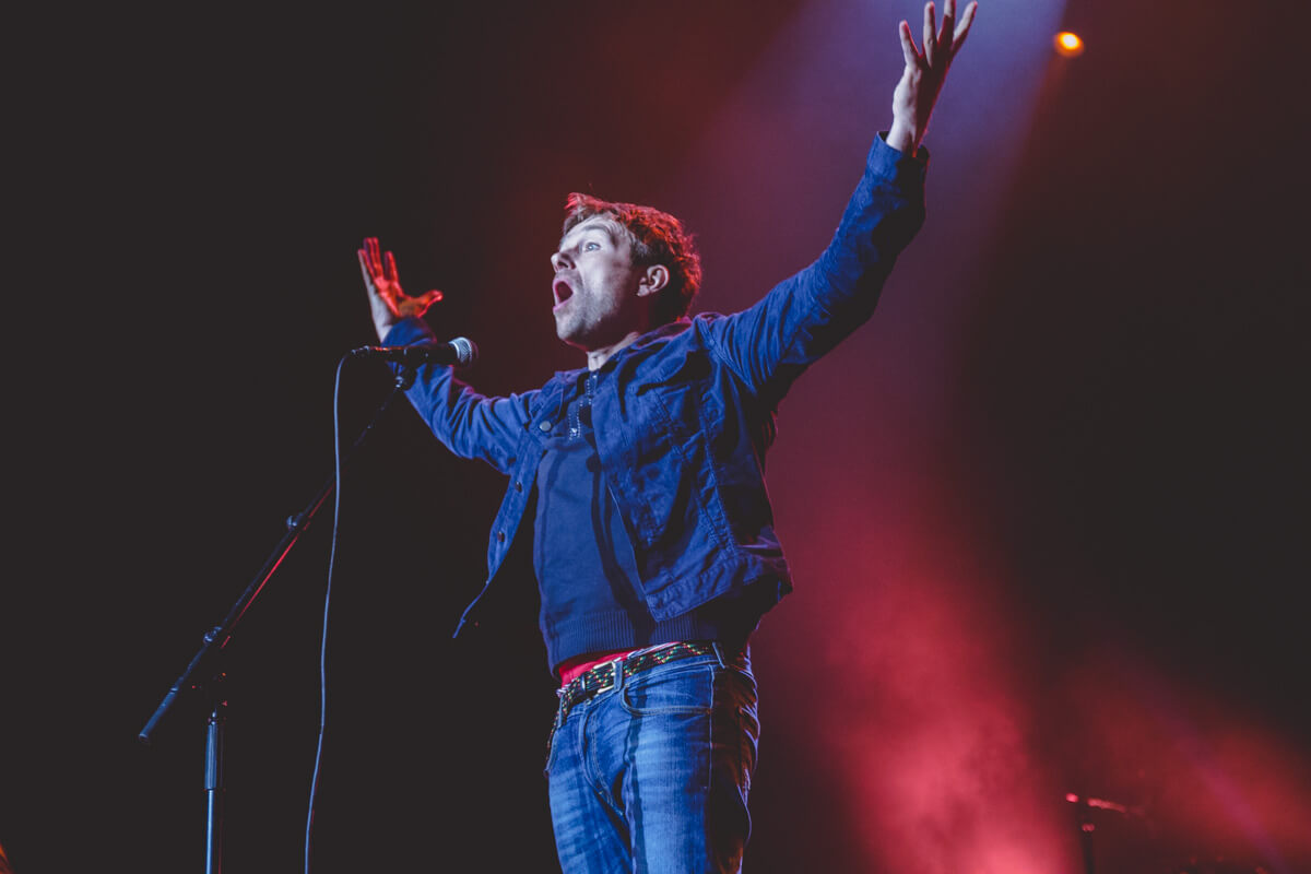 A man on stage singing, with his arms up in the air