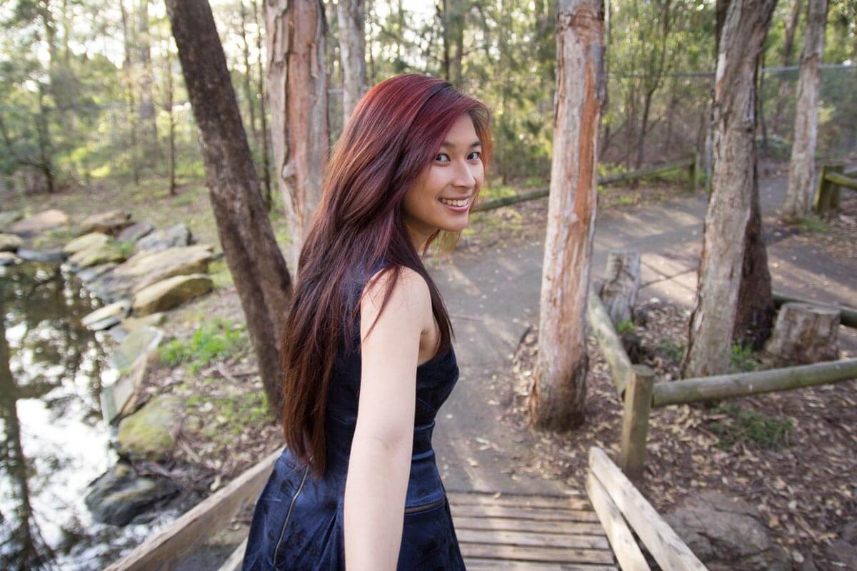 A woman with long red hair, wearing a blue dress, looking over her shoulder. She is in a park with a lot of trees in the background.